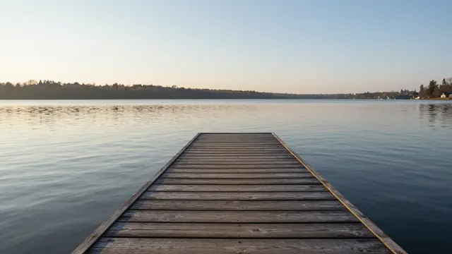 A wooden dock extends out over calm lake water toward a treeline under a soft sunrise sky