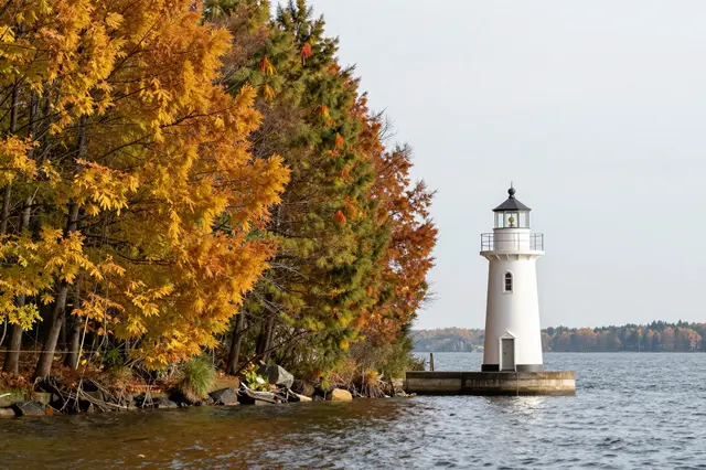 Small white lighthouse sits on a platform next to a shoreline of bright autumn trees on a calm lake