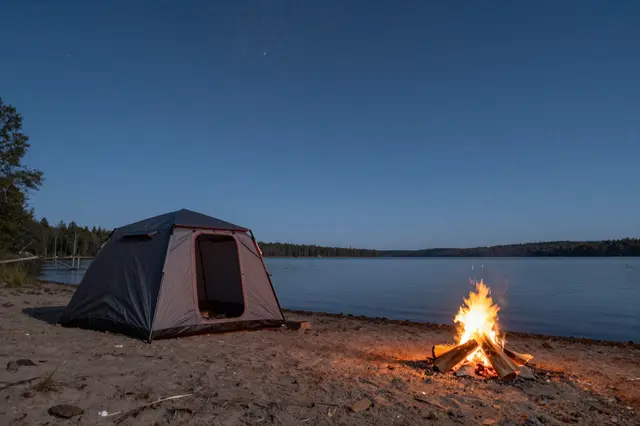 A tent sits on a sandy lakeshore next to a burning campfire under a clear starry evening sky