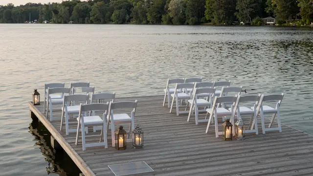 Empty white ceremony chairs and lit lanterns arranged on a wooden lakeside dock at dusk
