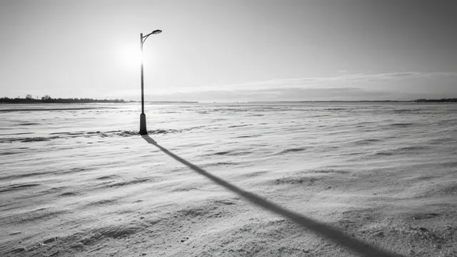 A lone lamppost casts a long straight shadow across wide fresh snow at low sunset in black and white