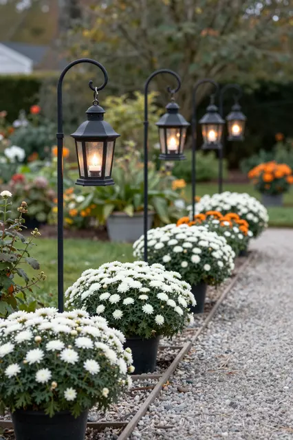 Lit black iron lanterns hang along a gravel path bordered with potted white and orange chrysanthemums