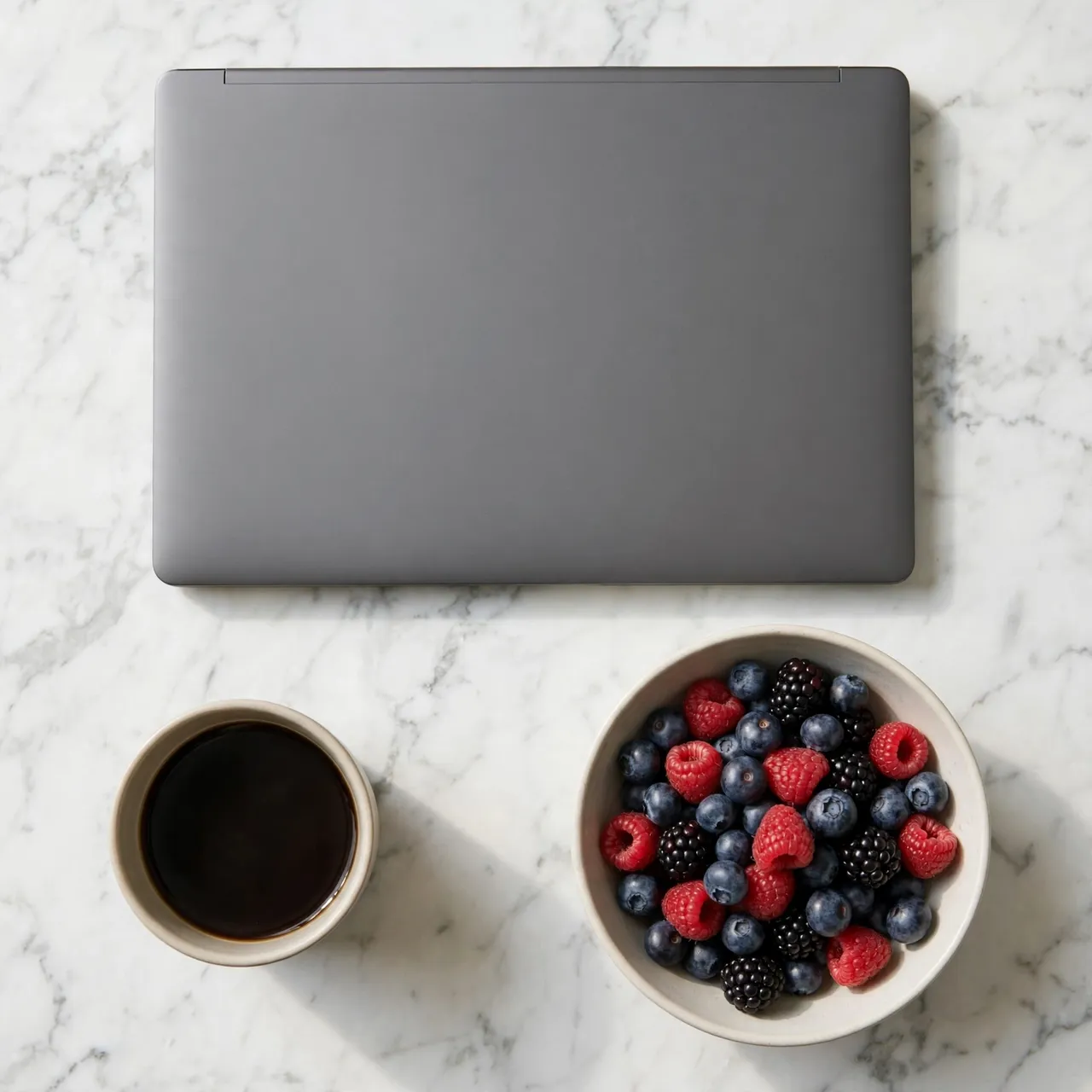 Overhead view of a closed gray laptop, coffee cup and berry bowl on white marble table