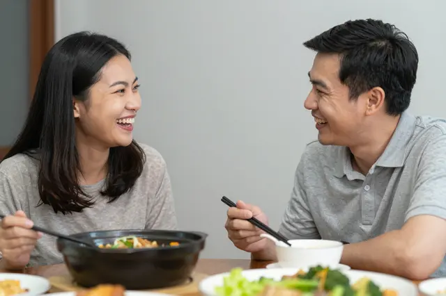 Two smiling adults laugh at each other while sharing a festive home dinner with chopsticks and shared dishes.