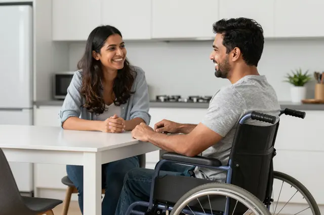 A smiling man in a wheelchair and a smiling woman sit facing each other across a bright kitchen table