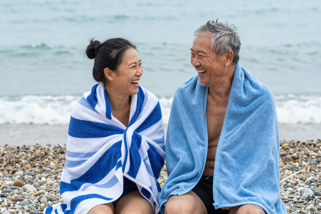 Two older people wrapped in towels laugh together on a pebble beach beside the sea