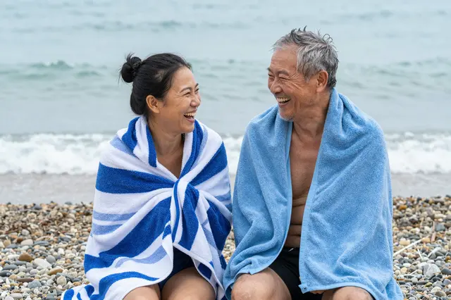 Two older people wrapped in towels laugh together on a pebble beach beside the sea