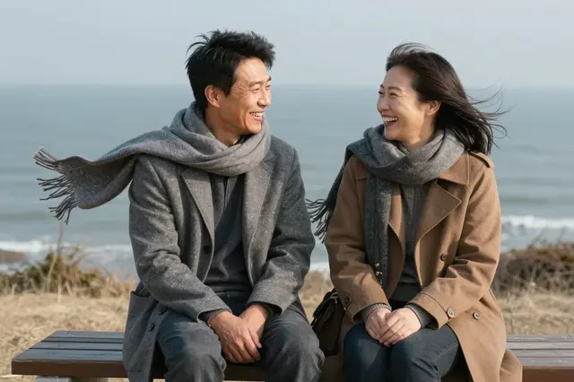 Two people in their forties laugh together sitting on a wooden bench at a windy coastal overlook