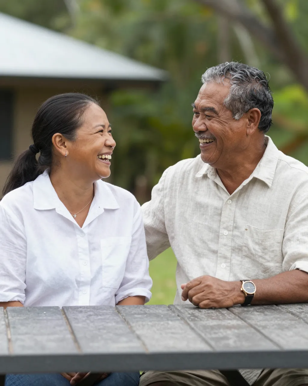 Pacific Islander senior man and adult woman smile and laugh at each other while sitting at a backyard wooden table