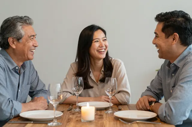 Three middle aged diverse friends laugh together around a candlelit wooden dinner table set with plates and wine glasses.