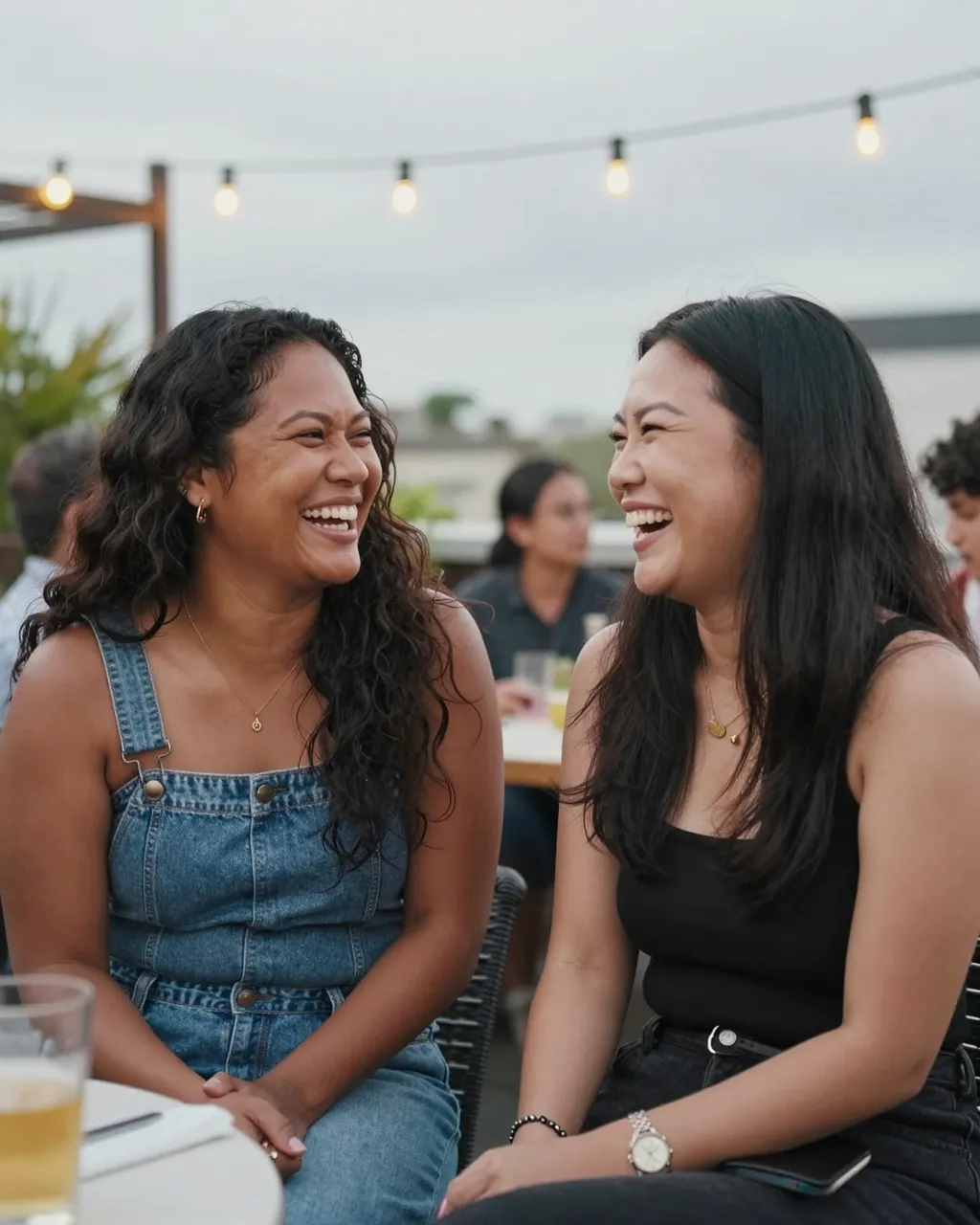 Two women friends laugh together while seated at an outdoor rooftop garden party under string lights