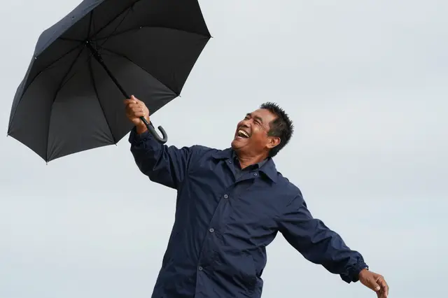A Pacific Islander man laughs while holding an umbrella caught in gusty wind under an overcast sky
