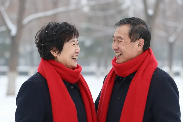 A senior East Asian couple wearing matching red scarves laugh while facing each other in a snow-covered park.