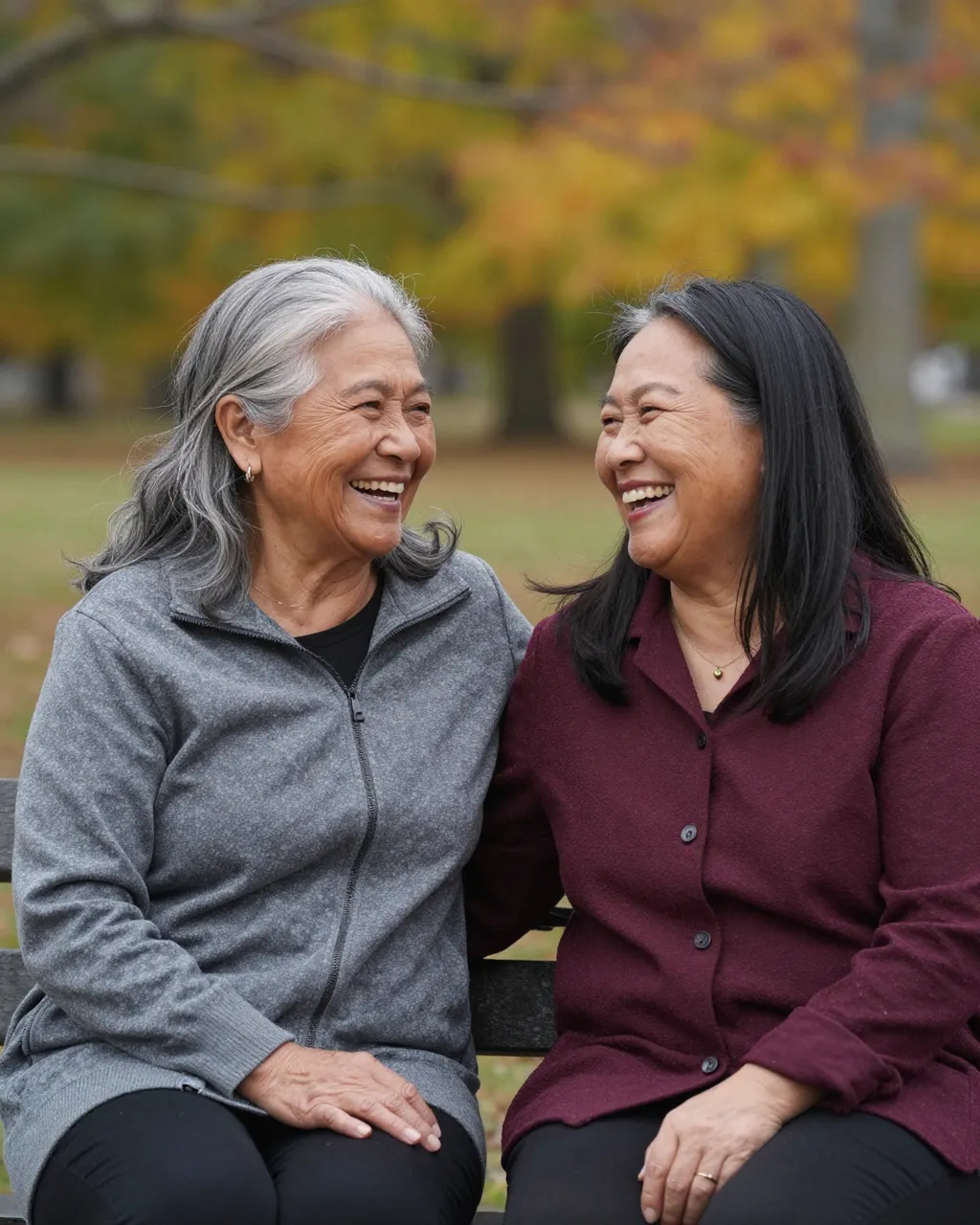 Two senior women laughing together while sitting shoulder to shoulder on an autumn park bench