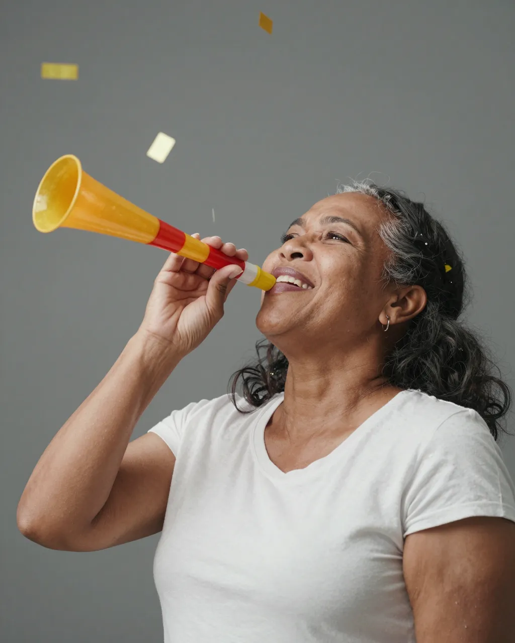 A smiling woman in her fifties blows a party horn as confetti falls around her