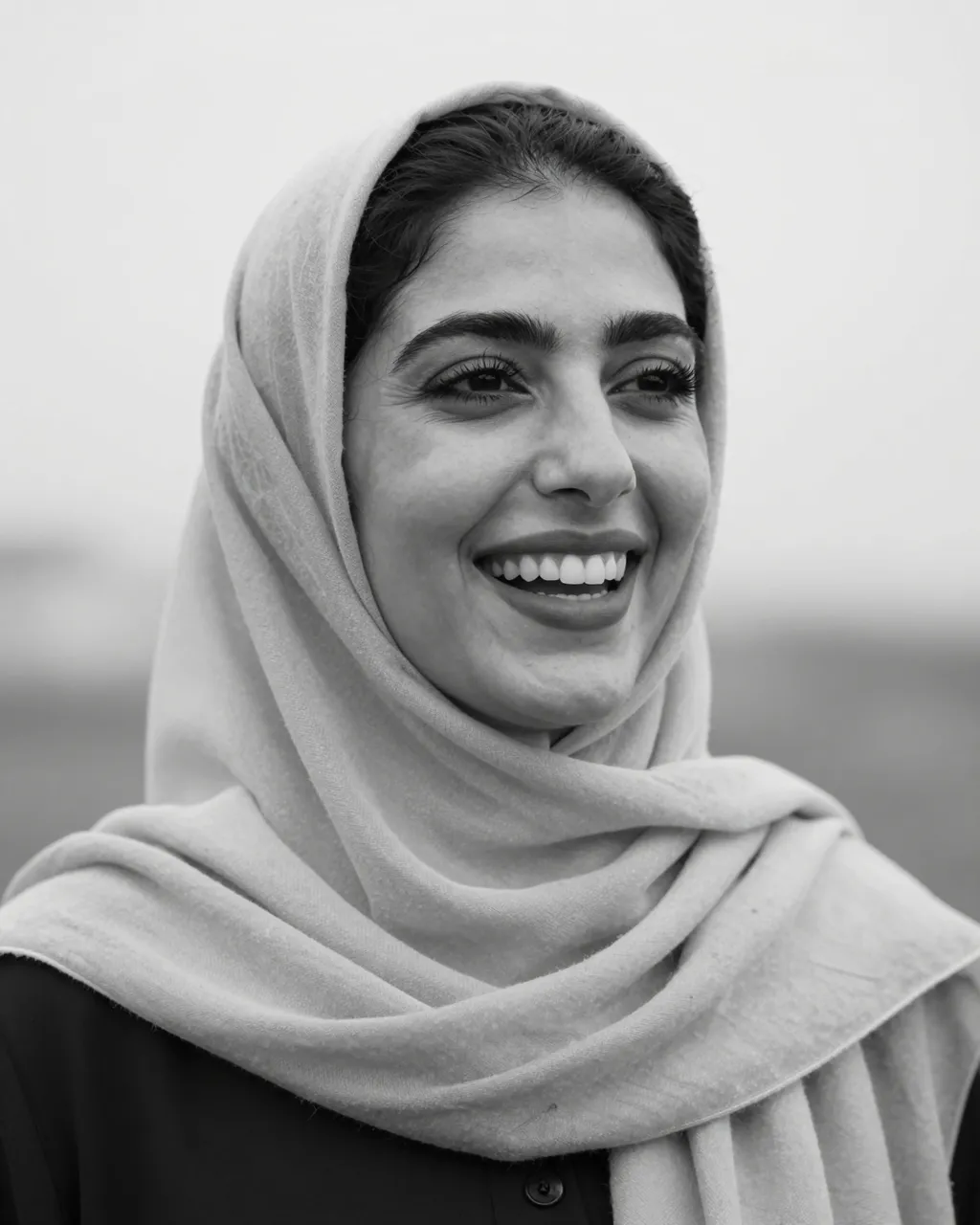 Black and white close-up portrait of a smiling Middle Eastern woman wearing a plain headscarf in soft overcast light.