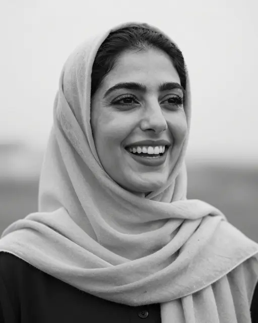 Black and white close-up portrait of a smiling Middle Eastern woman wearing a plain headscarf in soft overcast light.