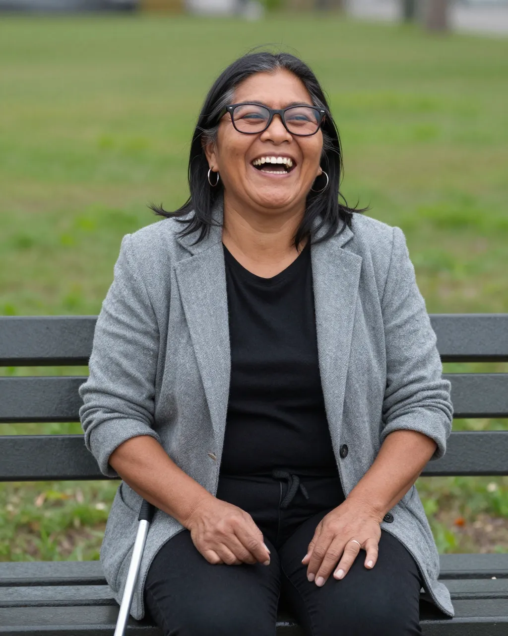 A laughing Indigenous woman sits on a park bench with a white cane beside her on grassy park grounds.