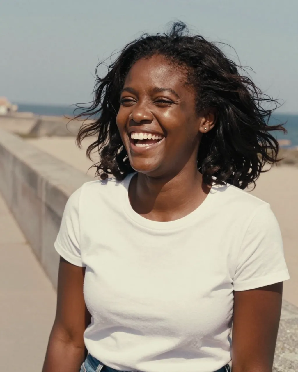 A Black woman in her late twenties laughs with wind blowing her hair while standing on a coastal seawall.