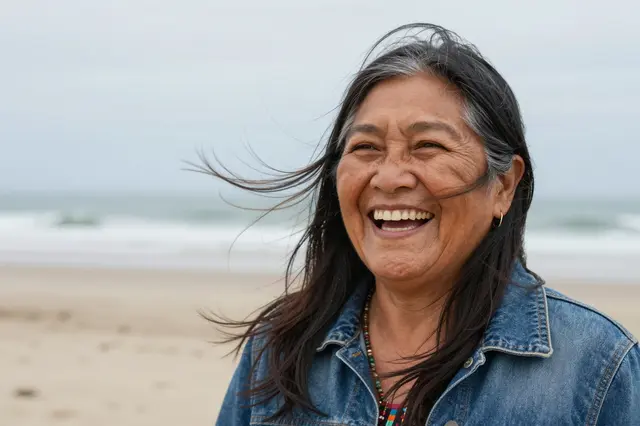 A smiling mature Indigenous woman stands on a beach with wind blowing her hair under an overcast sky.