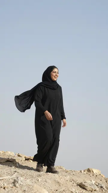 Smiling Middle Eastern woman with fluttering black hijab walks on a rocky cliff under a pale bright sky