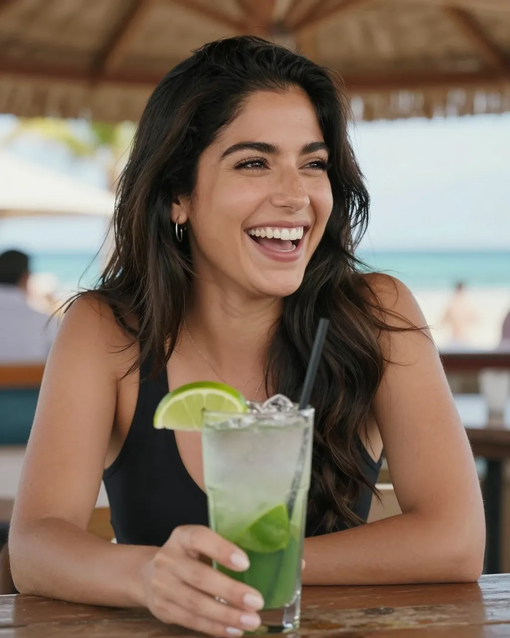 A smiling woman in her thirties holds a mojito at a sunny beachside bar under a palapa roof.