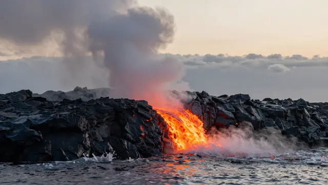 Glowing orange lava flows over dark volcanic rock into the ocean, creating steam plumes under a pale dusk sky
