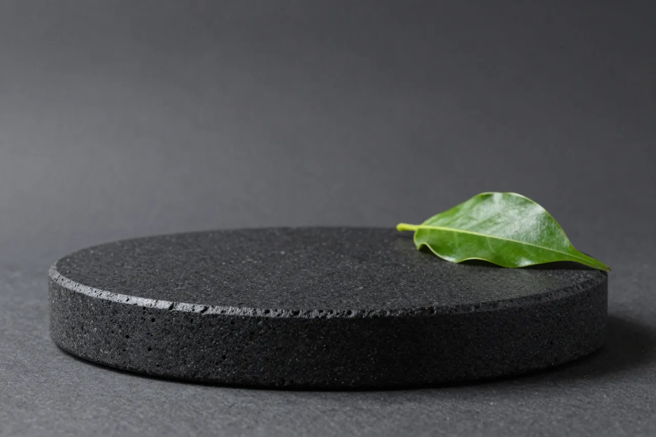 A round black porous lava stone product podium sits against a charcoal gray backdrop with a single green leaf resting on its