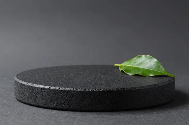 A round black porous lava stone product podium sits against a charcoal gray backdrop with a single green leaf resting on its