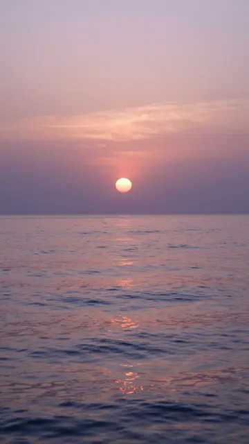 Round sun hangs over a calm rippled sea beneath a soft lavender and peach sunset sky