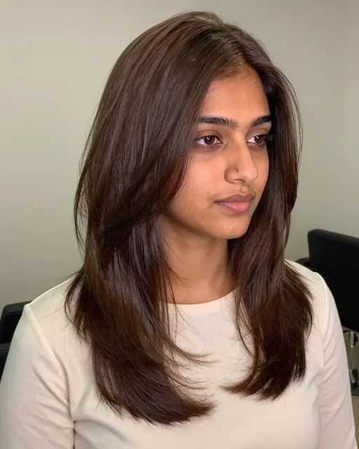 South Asian woman in her late twenties with glossy long layered brown hair wears an ivory top, looking off to the side