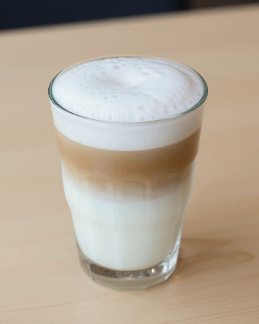 A tall clear glass of layered latte macchiato sits on a pale wooden table.