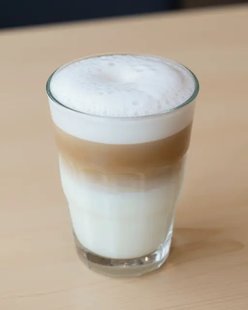 A tall clear glass of layered latte macchiato sits on a pale wooden table.