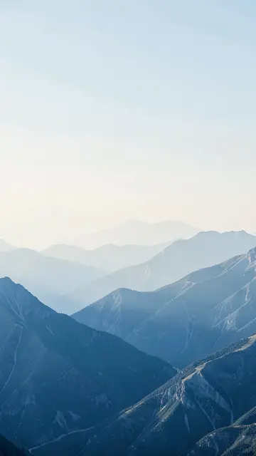 Vertical view of layered denim-blue mountain ranges receding under soft pale morning haze