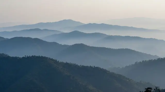 Stacked forested mountain ridges fade into cool blue mist under a pale hazy dawn sky