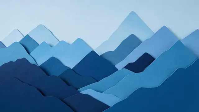 Flat lay layered paper cut mountain landscape in a gradient of blue tones against a pale sky background
