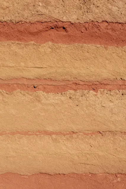 Full frame close-up of a rammed earth wall with horizontal layered bands of ochre, tan, and red clay.