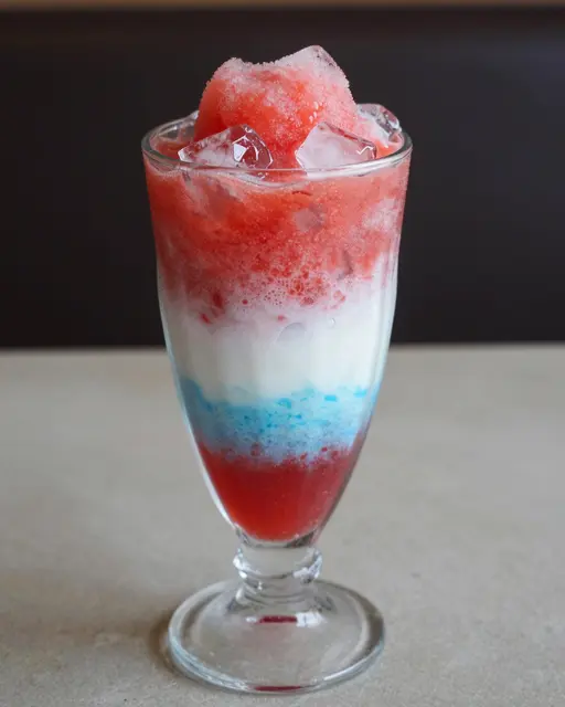 Tall glass holds a layered red, white, and blue slush drink topped with clear ice chunks.