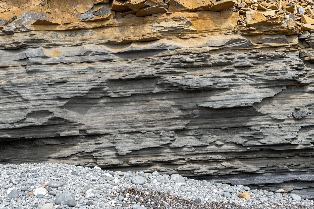 Eroded horizontal gray and ochre rock cliff rises above a pebble beach