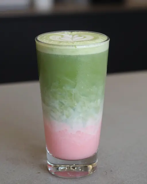 A tall glass holds a layered strawberry matcha latte with pink, milky and green layers and top latte art.