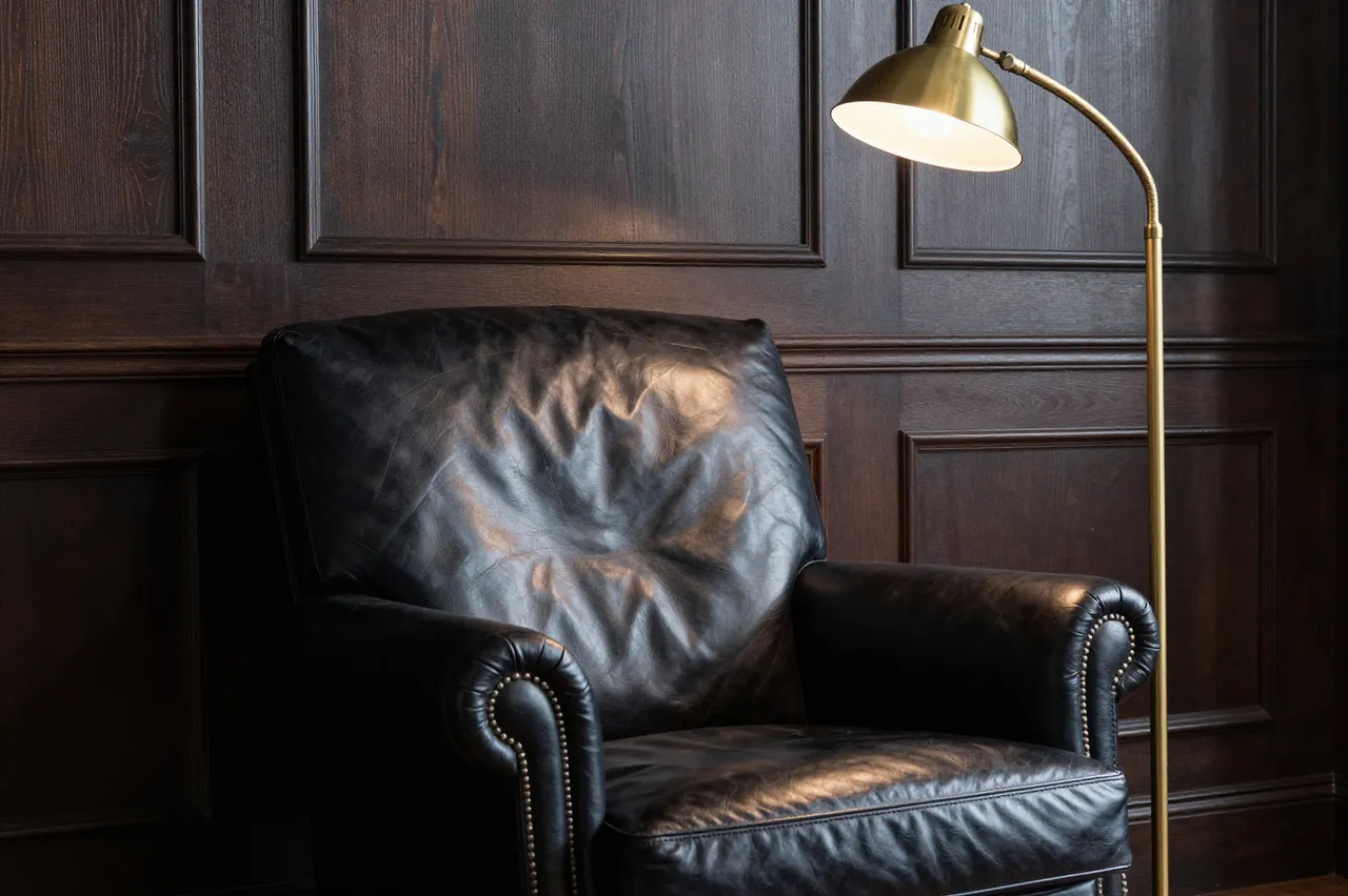 Lit brass floor lamp stands beside a studded black leather armchair against dark wood paneling