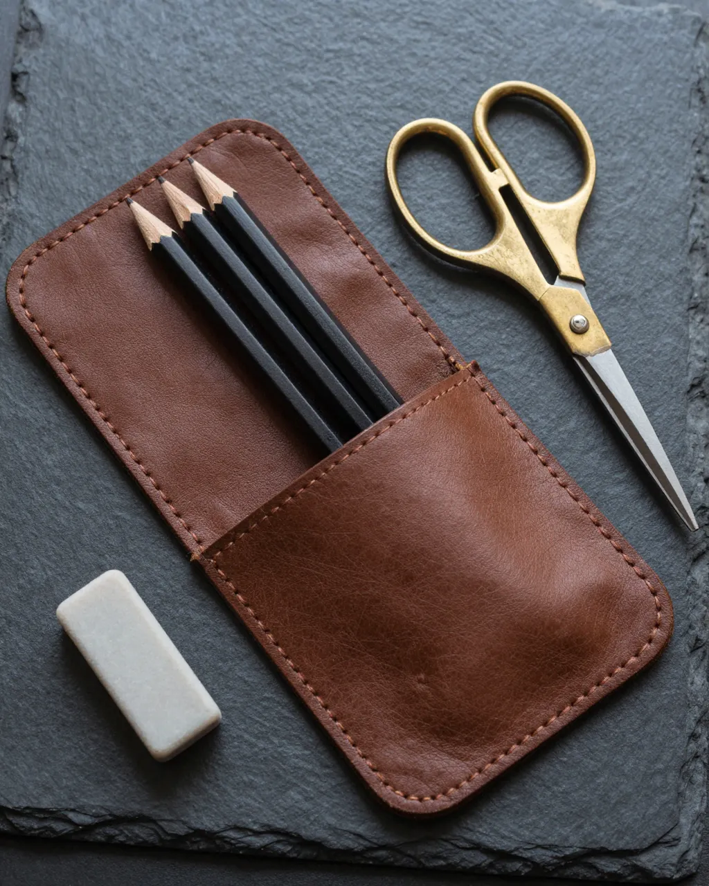 Flat lay of an open leather pencil roll, pencils, brass scissors and eraser on dark slate