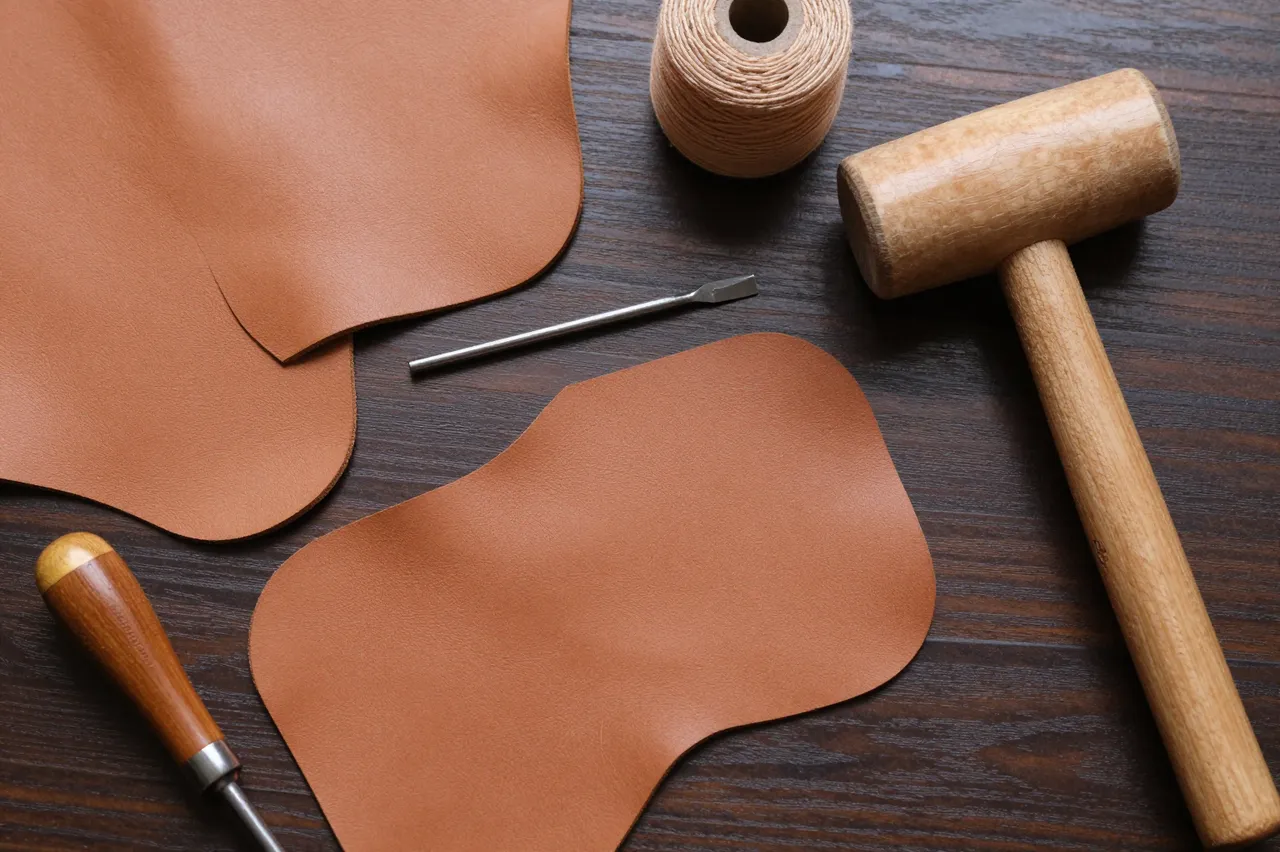 Flat lay arrangement of leatherworking tools and cut tan leather on a dark wooden work surface