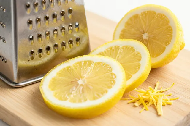 Sliced fresh lemon, zest, and a metal grater rest on a light wooden cutting board.