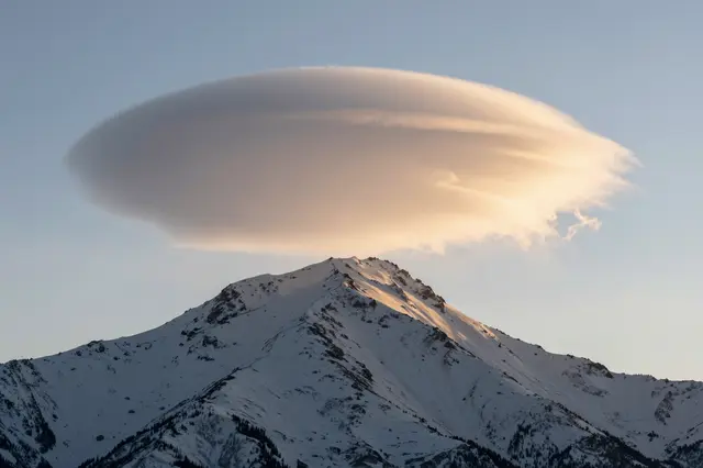 A large lens-shaped lenticular cloud hovers above a snow-covered mountain peak at golden sunset