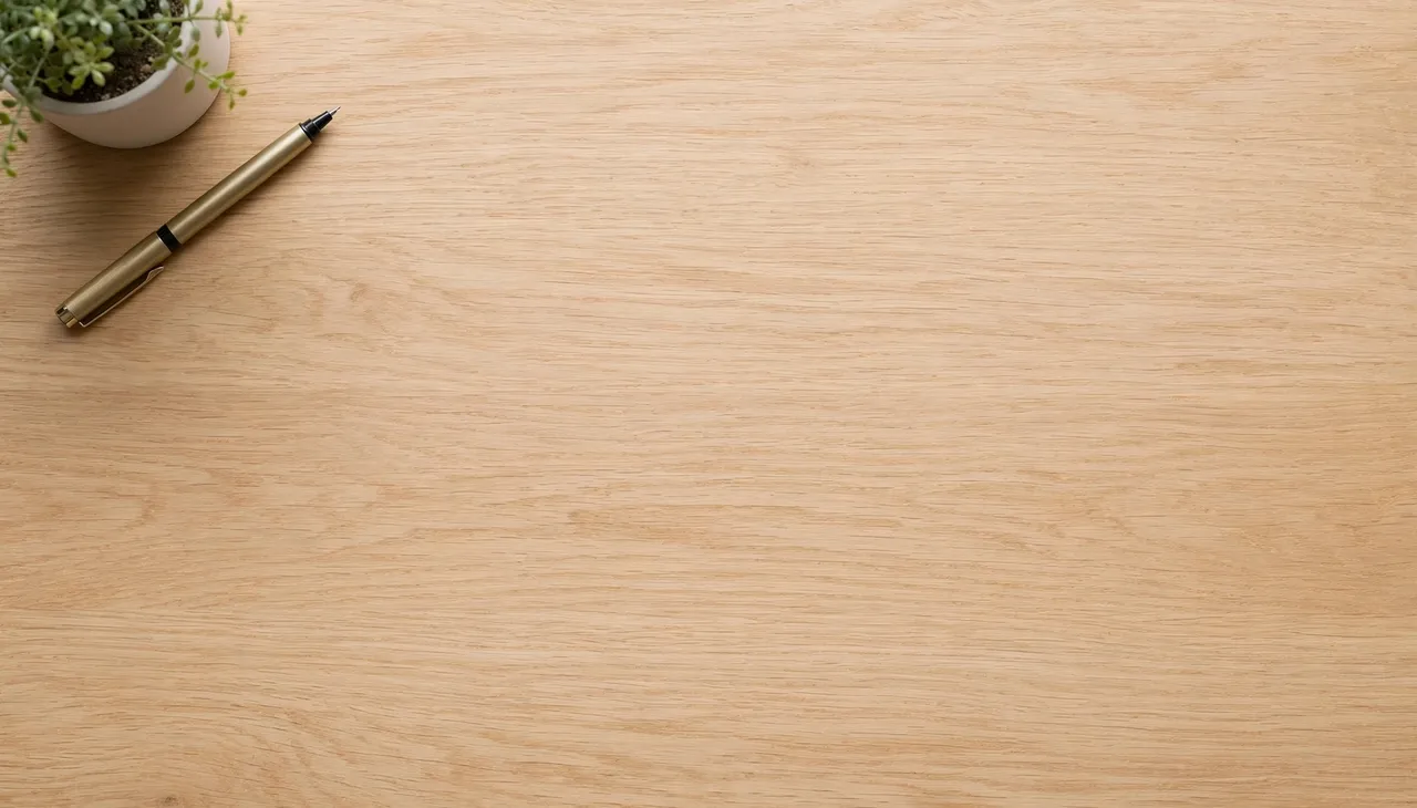 Overhead view of a light oak desk with a small potted plant and gold pen in the top left corner, leaving empty copy space.