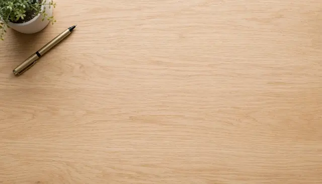 Overhead view of a light oak desk with a small potted plant and gold pen in the top left corner, leaving empty copy space.