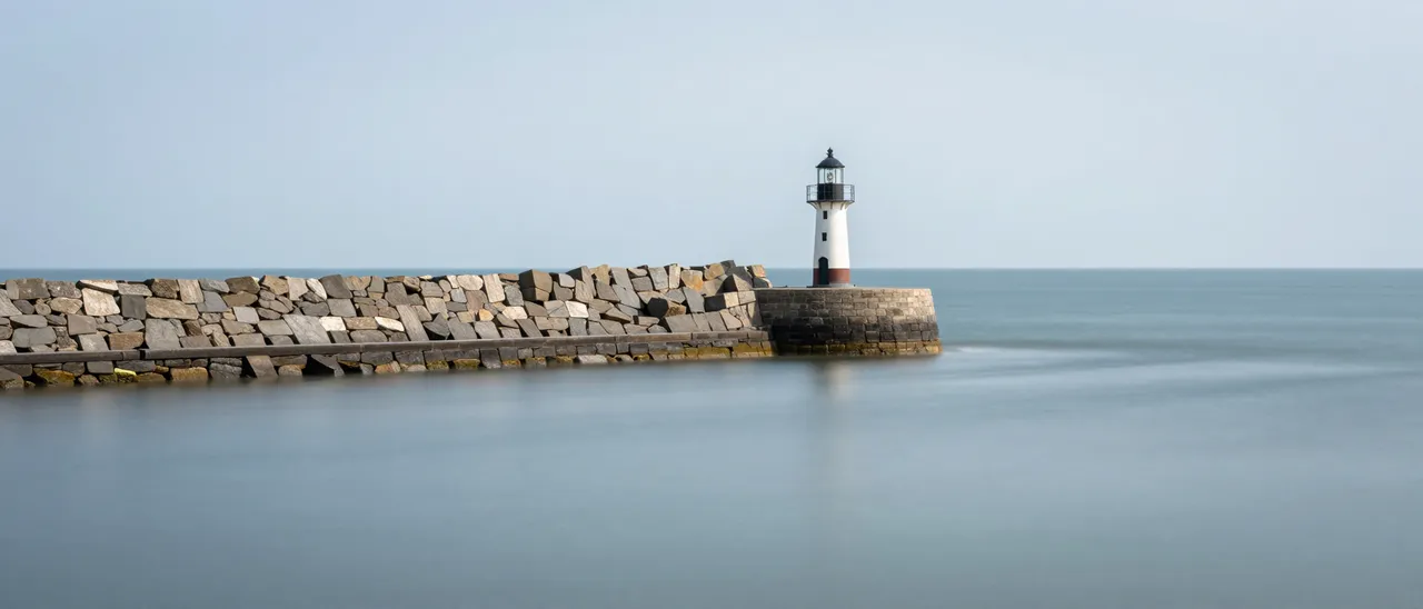 Long exposure view of a small lighthouse at the end of a stone breakwater over smooth glassy calm sea