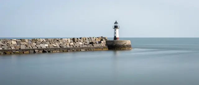 Long exposure view of a small lighthouse at the end of a stone breakwater over smooth glassy calm sea