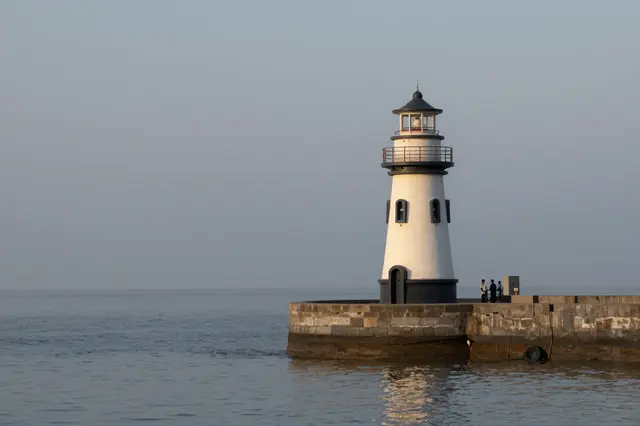 A white lighthouse stands on a stone pier overlooking calm hazy evening sea